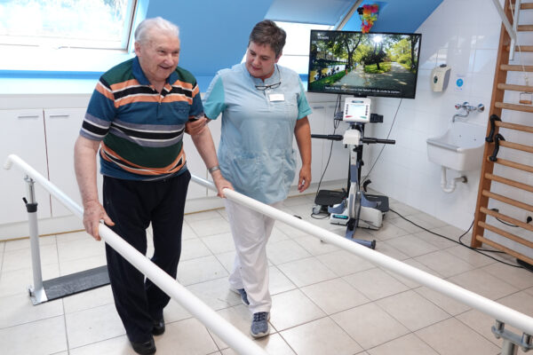 Een bewoner van woonzorgcentrum Coham voert stapoefeningen uit met behulp van leuningen en de begeleiding van een verpleegster in de kinesitherapieruimte. De ruimte is uitgerust met een turnrek, een lavabo en een hometrainer, die verbonden is met een televisie die een pad toont wanneer de trainer wordt gebruikt.