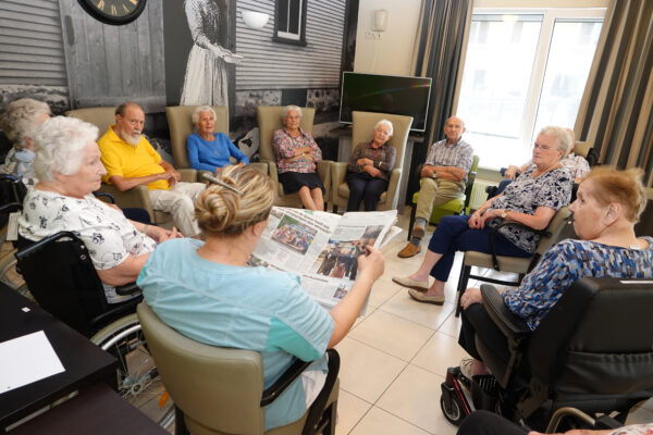 Een medewerkster van woonzorgcentrum Coham leest tijdens het krantenuur de krant voor aan tien bewoners. Vier bewoners zitten op een beige zetel, twee op een beige stoel en vier in hun persoonlijke rolstoelen. Ze zitten in een cirkel en luisteren aandachtig naar de medewerkster, die op een stoel zit.
