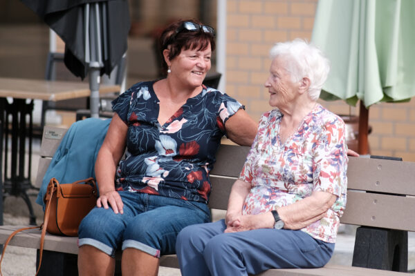 Een bewoonster van woonzorgcentrum Coham zit samen met haar bezoek op een bank op het terras van het woonzorgcentrum, waar ze een gesprek met elkaar voeren.