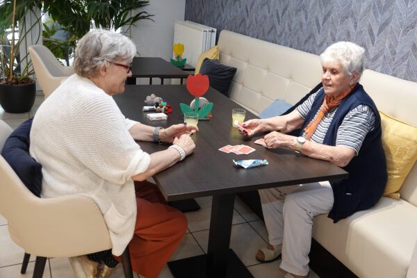 Twee bewoners van woonzorgcentrum Coham zitten samen aan een tafel en spelen een kaartspel. Ze drinken allebei een glas limonade, de één zit op een bank en de ander op een stoel.