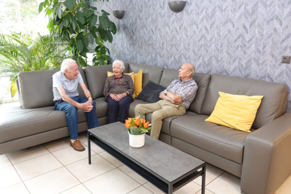 Drie bewoners van woonzorgcentrum Coham zitten samen op een grote hoeksofa in een zithoek. De sofa is versierd met drie gele kussens en een donkergrijs kussen.