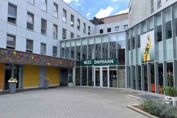 De ingang van woonzorgcentrum Damiaan in Tremelo. De gevel van de inkom is volledig van glas, terwijl de rest van het gebouw een grijskleurige gevel heeft. Boven de voordeur hangt een groot groen bord met witte letters die "WZC DAMIAAN" spellen. Direct aan de gevel is een abstracte versie van het logo te zien.