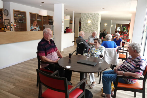De brasserie van woonzorgcentrum Damiaan. Op de voorgrond zitten twee groepen bewoners samen aan een tafel. De eerste groep bestaat uit drie personen: één drinkt een glas cola, een ander een glas water en de derde een glas wijn. De tweede groep bestaat uit vier personen, waarvan één in een rolstoel zit.