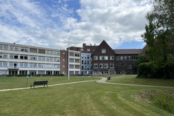 Het gebouw van woonzorgcentrum Damiaan in Tremelo, gezien vanuit de tuin. Het gras is net gemaaid en de tuin bevat meerdere paden, langs waarvan banken staan. Naast het gebouw is een terras met gesloten parasols. De gevel van het gebouw is opgebouwd uit verschillende materialen, zoals beton en baksteen.