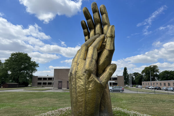 Een standbeeld van het logo van woonzorgcentrum Damiaan, die buiten die gelegen. Het standbeeld, in een gouden kleur, toont twee handen die elkaar verticaal vasthouden, waarbij de ene hand hoger is dan de andere.