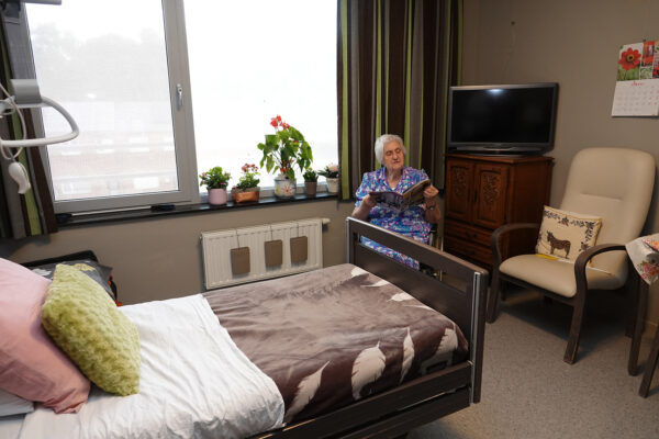 Een bewoonster zit in haar kamer in woonzorgcentrum Damiaan. De kamer is ingericht met een mooi opgemaakt bed, een antieke houten kast met daarop een tv, en een beige stoel met houten leuningen en poten. De vrouw zit in haar rolstoel in de hoek van de kamer en leest een tijdschrift.