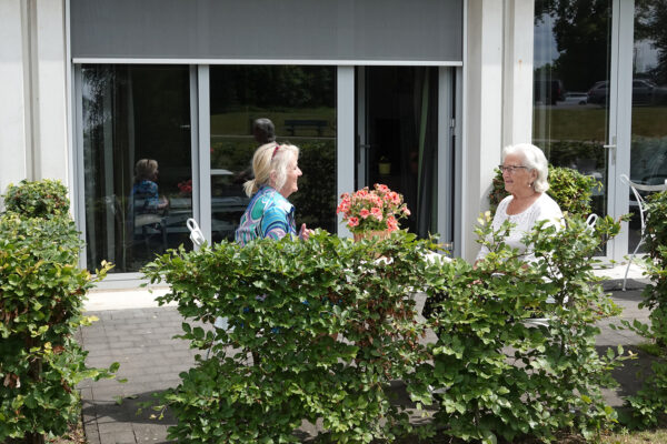 Twee bewoners van woonzorgcentrum Damiaan in Tremelo zitten aan een tafel op het terras te praten. Op de witte tafel staat een rieten vaas met roze bloemen. Achter hen is een deur te zien die direct naar de kamer van een van de bewoners leidt.