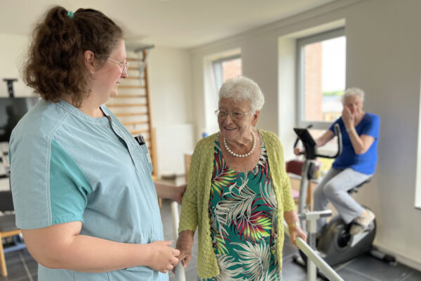 Een vrouwelijke bewoner doet wandeloefeningen in de kinesitherapieruimte van woonzorgcentrum Damiaan. Ze houdt zich vast aan de leuningen en wordt ondersteund door een verpleegster. Op de achtergrond is een mannelijke bewoner zichtbaar die aan het trainen is op een hometrainer.