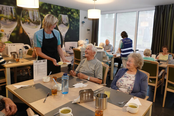 Een medewerkster van woonzorgcentrum Damiaan in Tremelo brengt eten naar de bewoners in het restaurant. Het restaurant is vol met mensen die net de soep als entree hebben gegeten. Het hoofdgerecht wordt nu geserveerd.