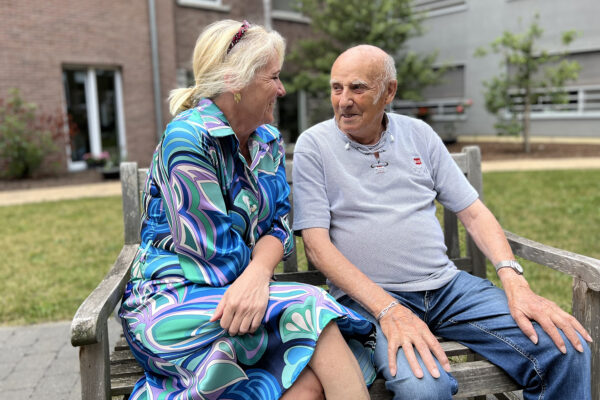 Twee personen zitten op een houten bank in de tuin van woonzorgcentrum Damiaan in Tremelo. Ze kijken elkaar aan en glimlachen. De vrouw draagt een jurk in verschillende tinten blauw, terwijl de man een jeans en een grijs T-shirt aan heeft.