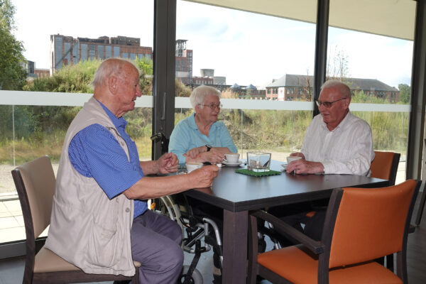 Drie bewoners zitten gezellig samen in de brasserie van woonzorgcentrum Sporenpark, genieten van een kopje koffie en voeren een aangenaam gesprek. Ze zitten naast grote ramen, die ook schuifdeuren zijn, waardoor er veel natuurlijk licht naar binnen stroomt.