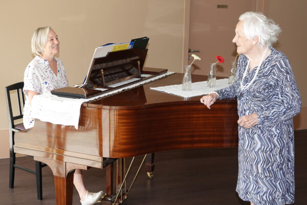 Een piano staat in de brasserie van woonzorgcentrum Sporenpark. Een vrijwilliger bespeelt de piano, terwijl een vrouwelijke bewoner rustig luistert naar de muziek terwijl ze naast de piano staat. Op de piano staan drie bloemen in glazen vazen.
