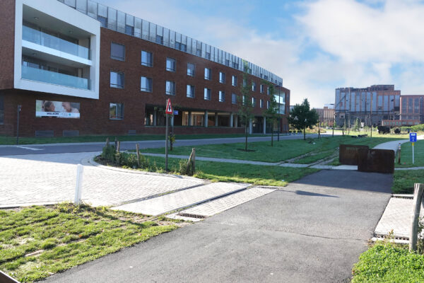 Straatzicht op het gebouw van woonzorgcentrum Sporenpark in Beringen, gelegen in een rustige straat met weinig verkeer. Een welkomstbord heet bezoekers welkom bij het woonzorgcentrum. De gevel is opgebouwd uit baksteen en wordt geaccentueerd door glazen en witte details bij de terrassen.