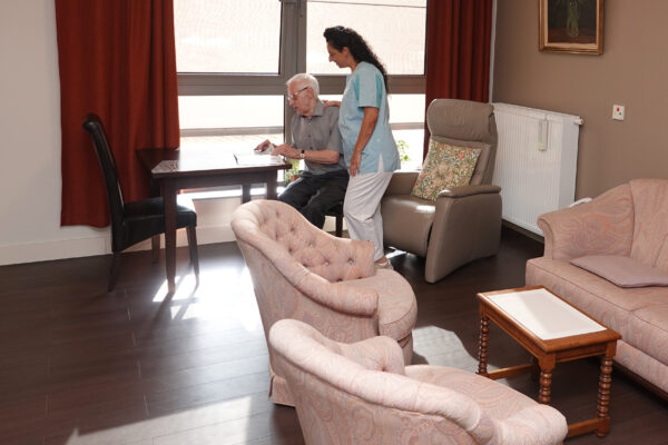 Een bewoner van woonzorgcentrum Sporenpark in Beringen zit in zijn kamer en toont iets aan een medewerkster die hem bezoekt. De kamer is ingericht met zijn persoonlijke meubels, waaronder twee roze zetels en een bijpassende sofa.