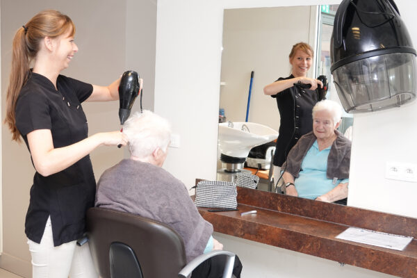De kapper droogt het haar van een bewoner van woonzorgcentrum Sporenpark in het kapsalon, nadat haar haar gewassen en geknipt is. Aan de muur hangt een spiegel.