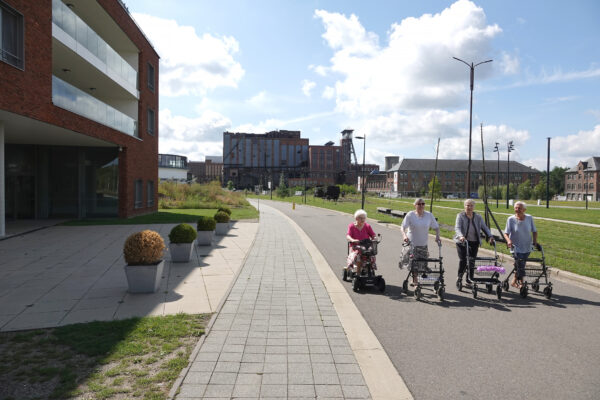 Vier bewoners maken een gezellige wandeling in de omgeving van woonzorgcentrum Sporenpark in Beringen. Het pad is breed en goed begaanbaar, waardoor de bewoners gemakkelijk hun rollators kunnen gebruiken en de route voor iedereen toegankelijk is.