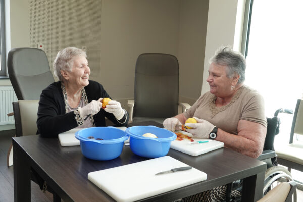 Twee bewoners van woonzorgcentrum Sporenpark in Beringen schillen samen appels in de ontspanningsruimte, terwijl ze gezellig met elkaar in gesprek zijn. Ze gebruiken handschoenen en een snijbord.