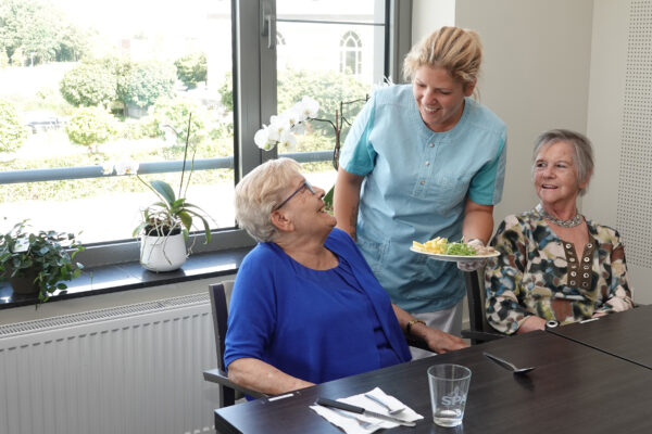Tijdens de maaltijdservice in woonzorgcentrum Sporenpark in Beringen serveert een medewerkster het eten aan twee bewoners die gezellig samen aan tafel zitten in het restaurant.