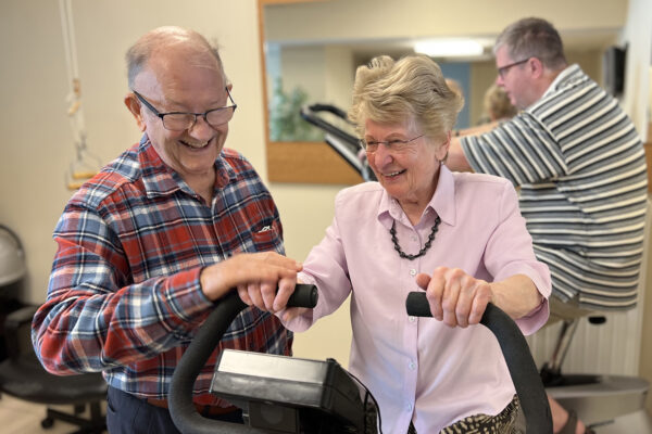 De gemeenschappelijke fitnessruimte op de campus van Ter Bleuk, die beschikbaar is voor alle bewoners van de assistentiewoningen. Een vrouw doet fietsoefeningen, terwijl haar man haar ondersteunt en helpt.