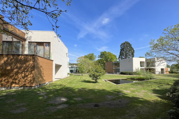 De buitenkant van de verschillende assistentiewoningen, waarbij duidelijk zichtbaar is dat elke woning beschikt over een privéterras met glazen leuningen. Daarnaast is er toegang tot de gemeenschappelijke tuin, die een klein vijvertje bevat. De gevels van het gebouw zijn opgebouwd uit witte bakstenen, terwijl de terrassen houten accenten hebben.