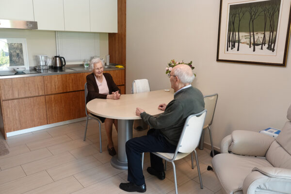Een man en vrouw zitten in de eetkamer van hun assistentiewoning en voeren een gesprek. De eetkamer is ingericht met een witte tafel en bijpassende stoelen. Op de voorgrond staat een witte fauteuil, terwijl op de achtergrond de keuken zichtbaar is, waar een oude familiefoto zorgvuldig is geplaatst.