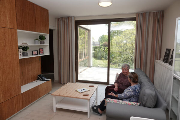 De woonkamer van een assistentiewoning op de campus van Ter Bleuk. Twee bewoners zitten gezellig op de sofa en voeren een gesprek. Tegen de muur staat een grote houten opbergkast met witte planken. Vanuit de woonkamer is er toegang tot een privéterras.