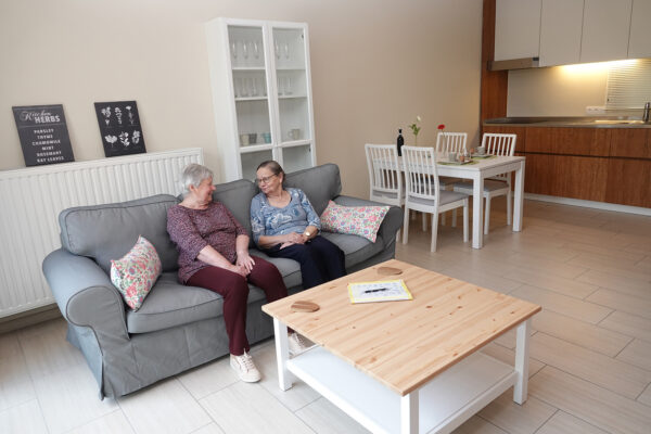 Twee bewoners van de assistentiewoningen Ter Bleuk zitten samen op een grijze sofa in de woonkamer van hun woning. In de ruimte staat een witte bijzettafel met een houten bovenblad en een witte vitrinekast, waarin glazen en bekers netjes zijn opgeborgen.