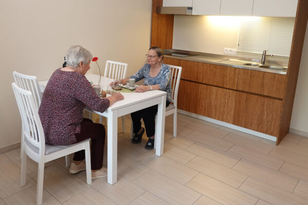 De eetkamer en keuken van een assistentiewoning op de campus van Ter Bleuk. Twee bewoners zitten aan een witte, rechthoekige tafel en drinken samen koffie terwijl ze gezellig met elkaar praten. In de keuken hebben de onderkasten een houtkleurige afwerking, terwijl de bovenkasten wit zijn.
