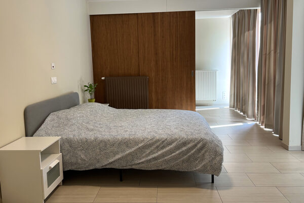 De slaapkamer van een assistentiewoning in Ter Bleuk. Een ruime, betegelde kamer met een dubbel bed en twee witte nachtkastjes. De kamer heeft een sfeervolle houten accentwand die de ruimte een warme uitstraling geeft.