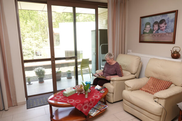 Een bewoner van een assistentiewoning in Ter Bleuk zit ontspannen in een van haar twee beige zetels in de woonkamer. Vanuit de woonkamer heeft ze toegang tot een privéterras. Aan de muur hangt een foto van haar kleinkinderen, terwijl ze zelf rustig een magazine leest.