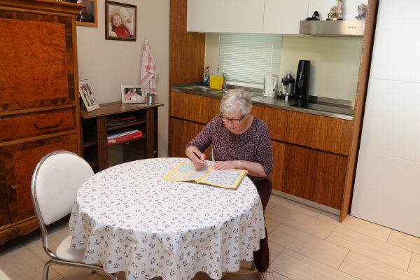 De eetkamer en keuken van een assistentiewoning in Ter Bleuk. De bewoner zit aan de tafel en maakt woordraadsels in een tijdschrift. Aan de muur hangt een foto van een baby, en op een bijzettafel staan familiefoto's van de bewoner. In de eetkamer staat een grote houten kast. De keuken is uitgerust met een dubbele kraan en een moderne kookplaat.