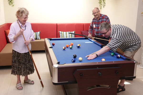 De ontspanningsruimte op de campus van de assistentiewoningen Ter Bleuk. In de ruimte staat een biljarttafel met een blauw biljartlaken en een grote roodbruine zetel. Twee bewoners spelen samen met hun familielid een potje biljart.
