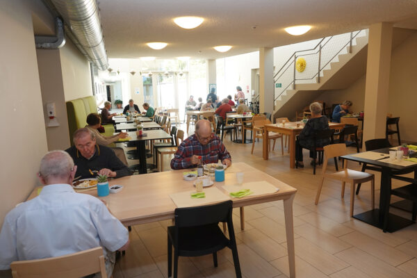 Het restaurant van de assistentiewoningen Ter Bleuk, waar bewoners samen aan een tafel zitten of alleen genieten van hun maaltijd. Aan één kant van het restaurant bevinden zich grote ramen die van het plafond tot de vloer reiken, waardoor er veel natuurlijk licht naar binnen stroomt.