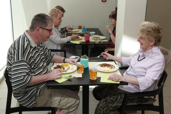 Het restaurant op de campus van de assistentiewoningen Ter Bleuk, toegankelijk voor alle bewoners. Op de voorgrond is een vrouwelijke bewoner te zien die samen met haar familielid van een maaltijd geniet. Op de achtergrond zitten drie andere bewoners samen en eten gezellig met elkaar.